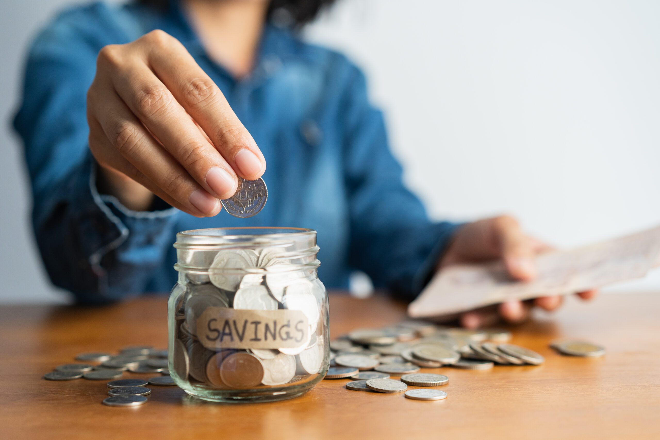  A person in a blue shirt is dropping a coin into a glass jar labeled "Savings" while holding a piece of paper that may be a bill.